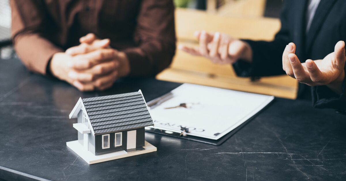 Two people discussing real estate investment strategy with a model house and contract on the table.